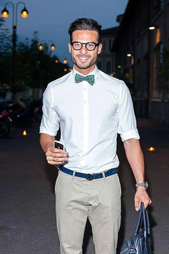 Hombre joven con gafas y pajarita verde con puntos blancos, camisa blanca y pantalones beige, en exterior nocturno