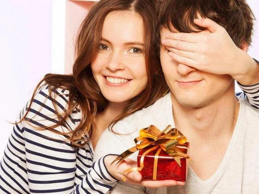 Pareja sonriente con regalo envuelto en caja roja y lazo dorado, mujer cubre ojos de hombre