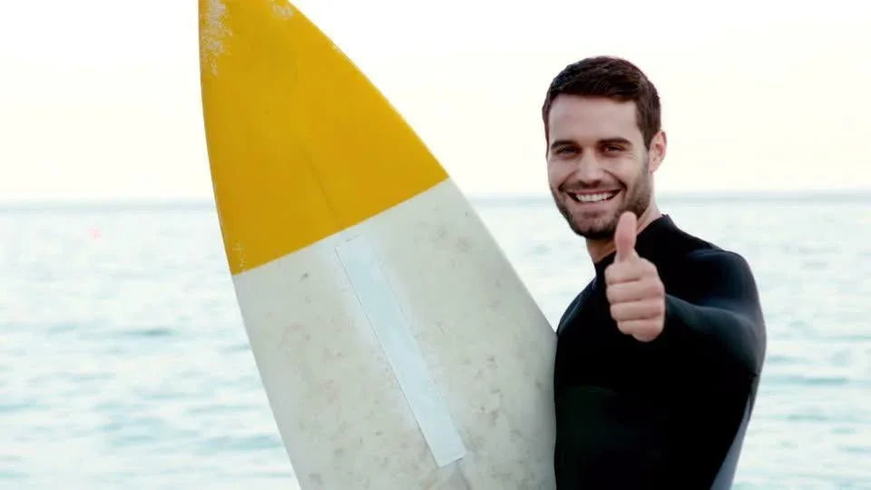 Hombre sonriente con traje de neopreno sostiene tabla de surf blanca y amarilla en la playa
