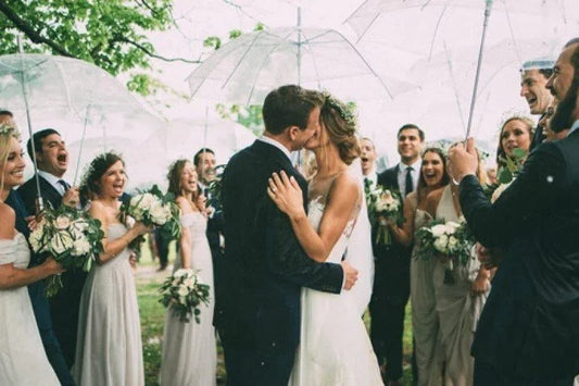 Novios besándose bajo paraguas transparentes con grupo de amigos en boda al aire libre vestidos elegantemente